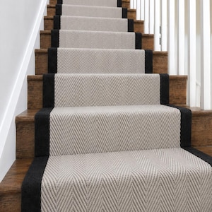 May include: A staircase runner featuring a light beige and white herringbone pattern with solid black fabric borders on each side. The runner is installed over wooden stair treads, extending from the bottom step up to the top of the staircase.