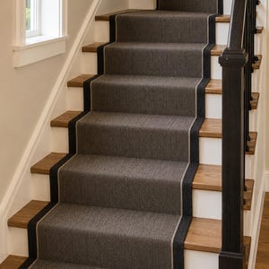 May include: A staircase featuring natural wood treads and white risers, covered by a dark gray woven stair runner. The runner has a thick black border with a thin light gray accent line running along the edges of the fabric on each step.
