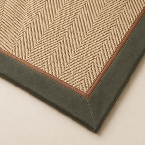 May include: A woven rug featuring a tan herringbone pattern in the center, bordered by a thin brown inner trim and a wide, dark olive green outer fabric border. The texture of the central weave is visible, showing a consistent, repeating zigzag design.