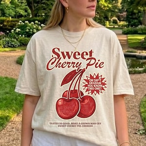 Puede incluir: Una camiseta de manga corta color crema con un gráfico de dos cerezas rojas y el texto Sweet Cherry Pie. El texto adicional dice OH SH*T GRAHAM YOU KNOW THIS ONE? y TASTES SO GOOD, MAKE A GROWN MAN CRY SWEET CHERRY PIE-EEEEEEE.