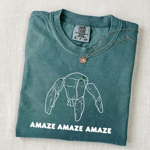 May include: A teal crew neck t-shirt with a white line art graphic of a robotic figure and the text "AMAZE AMAZE AMAZE" printed below. The shirt features a Comfort Colors brand label at the inner neckline and a thin gold chain necklace resting on top.