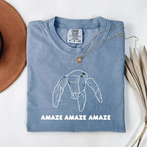 May include: A blue short-sleeved t-shirt featuring a white line-art graphic of a robot-like figure with small green accents. The text "AMAZE AMAZE AMAZE" is printed in white below the graphic. The shirt has a Comfort Colors brand label.