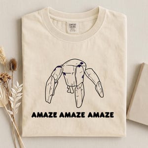 May include: A cream-colored short-sleeve t-shirt featuring a black line-art illustration of a blocky, multi-limbed robot figure centered on the chest. Below the graphic, the text "AMAZE AMAZE AMAZE" is printed in a bold, black sans-serif font.