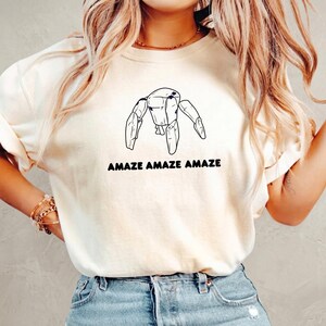 May include: A cream-colored short-sleeve t-shirt featuring a black line-art illustration of a robotic figure with three legs and the text "AMAZE AMAZE AMAZE" printed in a bold, rounded black font centered below the graphic design.