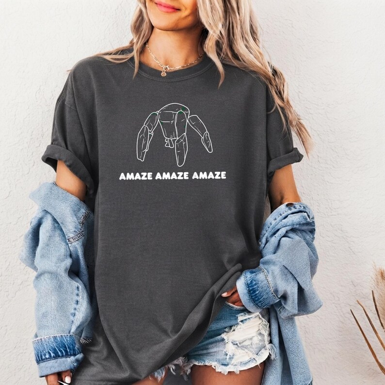 May include: A charcoal gray short-sleeve t-shirt featuring a white line-art graphic of a robotic figure with three legs and the text AMAZE AMAZE AMAZE printed in white capital letters centered below the illustration on the front of the garment.