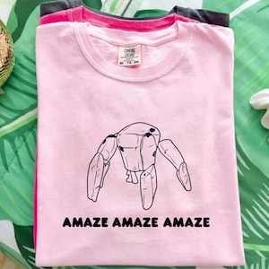 May include: A light pink short-sleeved t-shirt featuring a black line drawing of a robotic creature with four legs and the text "AMAZE AMAZE AMAZE" printed in black below the graphic. The shirt has a crew neckline and a Comfort Colors brand label.