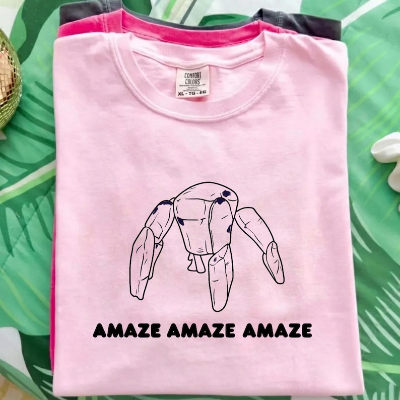 May include: A light pink short-sleeved t-shirt featuring a black line drawing of a robotic creature with four legs and the text "AMAZE AMAZE AMAZE" printed in black below the graphic. The shirt has a crew neckline and a Comfort Colors brand label.