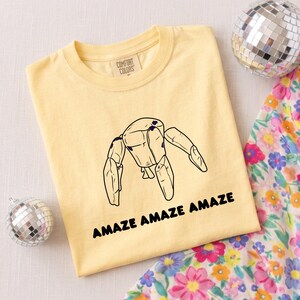 May include: A pale yellow t-shirt with a crew neckline and a Comfort Colors label. The center features a black line drawing of a jointed wooden mannequin figure with the text AMAZE AMAZE AMAZE printed in bold black capital letters below it.