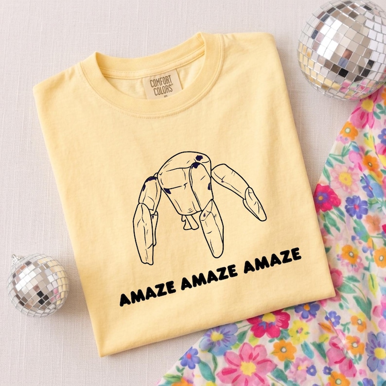 May include: A pale yellow t-shirt with a crew neckline and a Comfort Colors label. The center features a black line drawing of a jointed wooden mannequin figure with the text AMAZE AMAZE AMAZE printed in bold black capital letters below it.