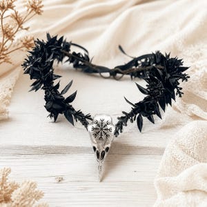 May include: A black floral crown featuring a silver-toned bird skull pendant at the centre. The skull is engraved with a black Vegvisir symbol. The crown is constructed from dark, pointed leaves and small blossoms arranged on a thin, flexible band.