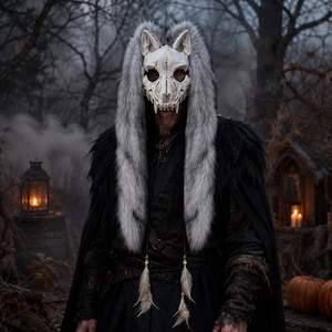 Op de afbeelding: Een wit dierenschedelmasker met puntige oren en scherpe tanden, omlijst door een lange, dikke grijs-witte imitatiebontrand. Het masker is bevestigd aan een zwarte capuchonmantel van donkere, gestructureerde stof met twee koorden met veren.