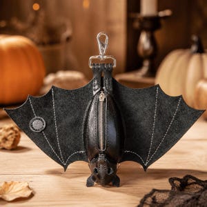 May include: A black leather bat-shaped coin purse featuring a central silver-toned metal zipper closure, stitched wing details, and a silver-toned swivel lobster clasp attached to the top for hanging. The design mimics a bat with outstretched wings.