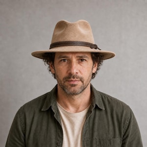 May include: A tan fedora hat featuring a dark brown leather band with a small knot detail. The hat has a structured crown with a central crease and a wide, flat brim. The material appears smooth and uniform in colour, providing a classic look.