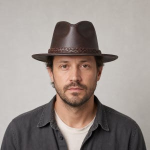 Peut inclure: Un chapeau fedora marron foncé doté d'une calotte pincée et d'un large bord. Le chapeau est orné d'une bande en cuir tressé qui entoure la base de la calotte, offrant un détail texturé contrastant avec la finition lisse du matériau.