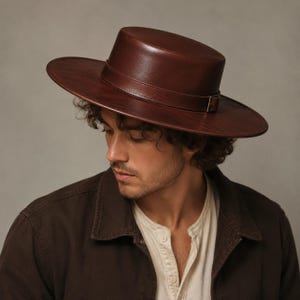 Puede incluir: Un sombrero de color marr&oacute;n oscuro con la parte superior plana, ala ancha y r&iacute;gida, y una banda a juego con una peque&ntilde;a hebilla met&aacute;lica. El sombrero tiene un acabado liso y estructurado con una silueta circular limpia y bien definida.