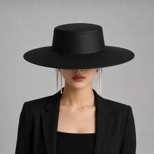 May include: A black wide-brimmed hat with a flat crown and a structured silhouette. The hat is paired with long, dangling earrings featuring multiple small white beads arranged in a vertical line, worn with a black blazer and a black top.