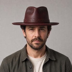 Op de afbeelding: Een diepbordeauxrode fedora-hoed met een gestructureerde kroon en een brede, platte rand. De hoed is voorzien van een bijpassende leren band rond de basis van de kroon, gedetailleerd met een dubbele rij kleine, gelijkmatig verdeelde gaatjes.