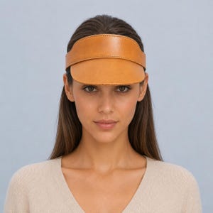 Könnte beinhalten: Ein beigefarbener Lederschirm mit glatter, matter Oberfläche und einem strukturierten, geschwungenen Schild. Der Schirm weist sichtbare Nähte entlang der Kanten des Stirnbandes und des Schildes auf, was für ein sauberes, einheitliches Aussehen sorgt.
