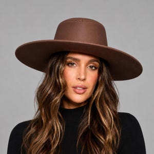 May include: A wide-brimmed brown hat with a structured crown and a flat top. The hat features a smooth, solid-colored surface and a clean, circular brim that extends outward, providing a consistent silhouette around the entire head area.