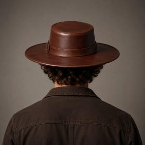 Puede incluir: Un sombrero de color marr&oacute;n oscuro con parte superior plana, ala ancha y una banda de cuero sint&eacute;tico a juego con una peque&ntilde;a hebilla met&aacute;lica. El sombrero se lleva sobre cabello oscuro rizado y una chaqueta de cuello marr&oacute;n oscuro, vista trasera.