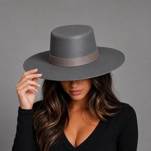 May include: A wide-brimmed hat in a solid matte grey finish featuring a flat crown and a structured circular brim. The hat is accented with a smooth, slightly darker grey band encircling the base of the crown, providing a clean and uniform look.