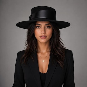 May include: A black wide-brimmed fedora hat featuring a smooth, satin-finish black ribbon band wrapped around the crown. The hat is worn with a black blazer and a delicate gold necklace with a small circular pendant resting on the chest.