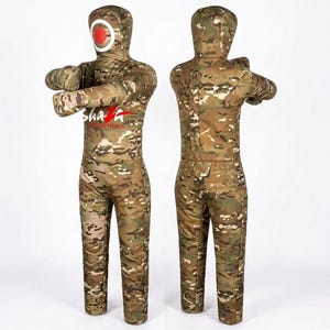 May include: Two human-shaped grappling dummies featuring a camouflage pattern in shades of brown, green, and tan. The front dummy displays a red and white circular target on the face and the text Shaza Industries on the chest area.