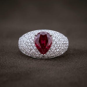 Könnte beinhalten: Ein silberfarbener Ring mit einem großen, birnenförmigen, tiefroten Edelstein in der Mitte, der von drei Krappen gehalten wird. Die breite Ringschiene ist dicht mit zahlreichen kleinen, klaren, rund geschliffenen Steinen besetzt.