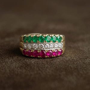 Puede incluir: Un anillo de oro con tres filas horizontales de piedras preciosas redondas facetadas. La fila superior presenta esmeraldas de color verde vibrante, la fila central contiene diamantes brillantes y la fila inferior muestra rubíes de color rojo intenso.