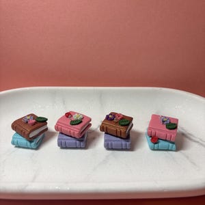 May include: Four miniature book charms arranged in pairs, each featuring a top book decorated with small sculpted flowers and leaves. The books are rendered in brown, pink, and light blue, with each pair stacked vertically on a white marble tray.
