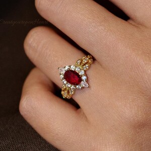 May include: A gold ring featuring a central oval-cut deep red gemstone surrounded by a halo of small clear stones. The band has an intricate, textured leaf-like design on both sides, extending from the central setting to create a detailed look.