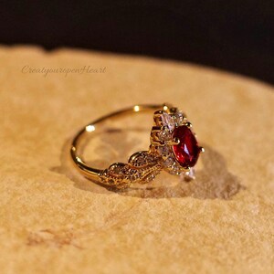 May include: A gold-toned ring featuring a central oval-cut red gemstone surrounded by a halo of small clear stones. The band is designed with a leaf-like motif encrusted with tiny clear stones. The text CreatyouropenHeart is visible.
