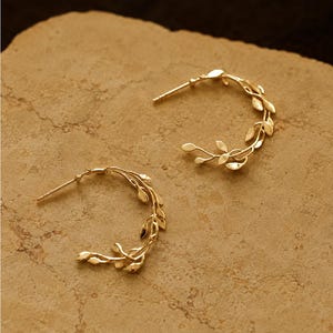 May include: A pair of gold hoop earrings featuring a delicate vine design with small, polished leaves attached to the circular frame. Each earring includes a thin post and a secure backing mechanism, showcasing a detailed botanical metalwork pattern.