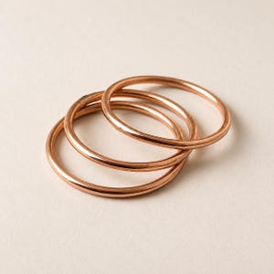 May include: Three thin, smooth, circular rings with a polished rose gold finish. The rings are stacked slightly overlapping each other, showcasing a simple, unadorned design with a reflective metallic surface and a consistent, rounded band profile.