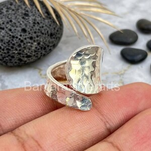 Könnte beinhalten: Ein silberfarbener Ring mit einer breiten, offenen Ringschiene und einer gehämmerten, texturierten Oberfläche. Das Design besteht aus einem großen, abgerundeten, konkaven Blickfang, der sich um den Finger legt und ein glänzendes Finish aufweist.