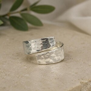 Puede incluir: Un anillo envolvente de tono plateado con una superficie texturizada y martillada. La banda está abierta en la parte delantera, con dos extremos superpuestos que crean un diseño cruzado. El metal tiene un acabado reflectante con hendiduras.