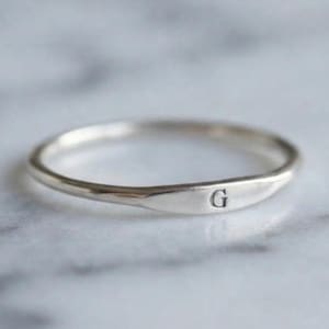 May include: A thin, polished silver-tone band ring featuring a small, flat, horizontal bar section on the front. The flat bar is engraved with the uppercase letter G in a simple, clean serif font. The ring has a smooth, reflective metallic finish.