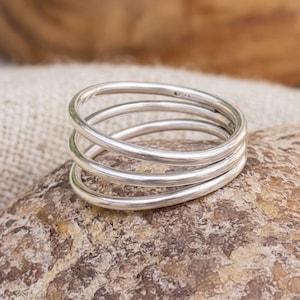 Puede incluir: Un anillo de plata con un diseño de múltiples bandas compuesto por tres bucles finos, lisos y continuos que se superponen para crear una apariencia en capas. El metal tiene un acabado pulido y reflectante con un perfil redondeado y limpio.