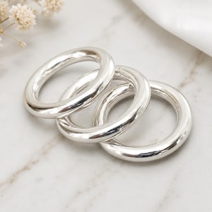 Puede incluir: Tres anillos de metal plateado pulido con una superficie lisa y redondeada. Los anillos están dispuestos en una formación superpuesta y entrelazada, mostrando un acabado reflectante y brillante que capta la luz en sus bandas circulares.