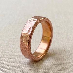 Pode incluir: Um anel de cobre maciço com uma textura martelada distinta em toda a sua superfície exterior. A banda tem um tom castanho-avermelhado metálico quente com um acabamento polido que reflete a luz, realçando as irregularidades da peça.