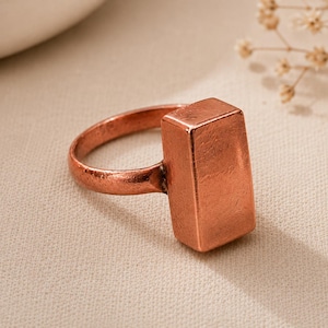 Peut inclure: Une bague de couleur cuivre dotée d'un bloc rectangulaire lisse en guise de pièce centrale, fixée sur un anneau circulaire. La surface métallique présente une finition texturée et légèrement patinée avec des variations de couleur cuivre.