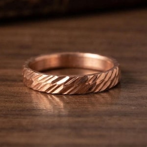 Peut inclure: Une bague en or rose présentant une surface texturée avec des rainures diagonales répétitives gravées sur l'anneau en métal. La bague a une largeur constante et une finition polie qui reflète la lumière sur le motif gravé et incliné.