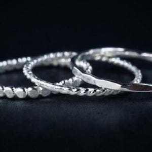 Può includere: Un set di tre anelli color argento disposti in fila. Il primo anello presenta una texture a perline, il secondo ha un design a corda intrecciata e il terzo mostra una superficie liscia e martellata con riflessi metallici brillanti.