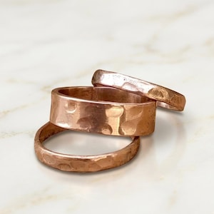 May include: A set of three copper-colored rings with a hammered, textured finish. The rings vary in width, with one wider band positioned between two thinner bands, all displaying a metallic, reddish-brown surface with visible indentations.