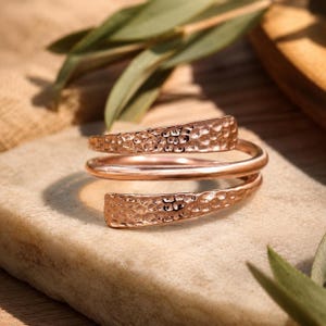 Puede incluir: Un anillo de oro rosa con diseño bypass que presenta una banda central lisa y pulida, flanqueada por dos extremos cónicos con un acabado martillado. El metal tiene un brillo cálido y el diseño se envuelve para crear una silueta abierta ajustable.