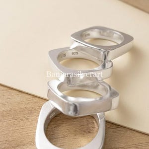 Può includere: Una pila di quattro anelli in argento sterling lucido con un design a fascia di forma quadrata. Ogni anello presenta una superficie liscia e riflettente ed è marchiato con il punzone 925 all'interno, che indica la purezza del metallo.
