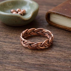 Può includere: Un anello in rame con un design a filo intrecciato. La fascia è composta da più fili di rame metallico intrecciati insieme in un motivo continuo, creando una forma circolare strutturata con una calda finitura metallica color bruno-rossastro.