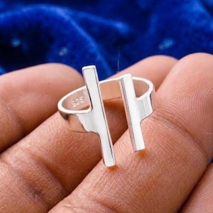 Puede incluir: Un anillo abierto de plata con dos elementos de barra vertical que se extienden desde la banda. El interior de la banda está claramente marcado con el sello 925, que indica la composición del metal. El anillo tiene un acabado pulido y liso.