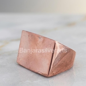 Può includere: Un anello in rame massiccio caratterizzato da un'ampia faccia rettangolare piatta con una finitura martellata e testurizzata. Il metallo ha una calda lucentezza metallica bruno-rossastra e un design a fascia spessa. Il testo Banjarasilverarts è visibile.