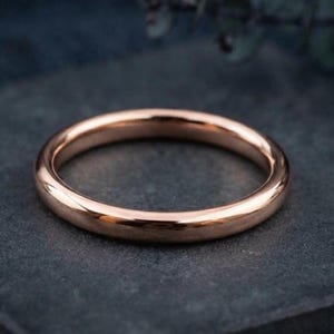 May include: A polished rose gold band ring with a smooth, rounded surface and a continuous circular shape. The metal has a warm, metallic pink hue that reflects light evenly across its simple, unadorned, and minimalist design.